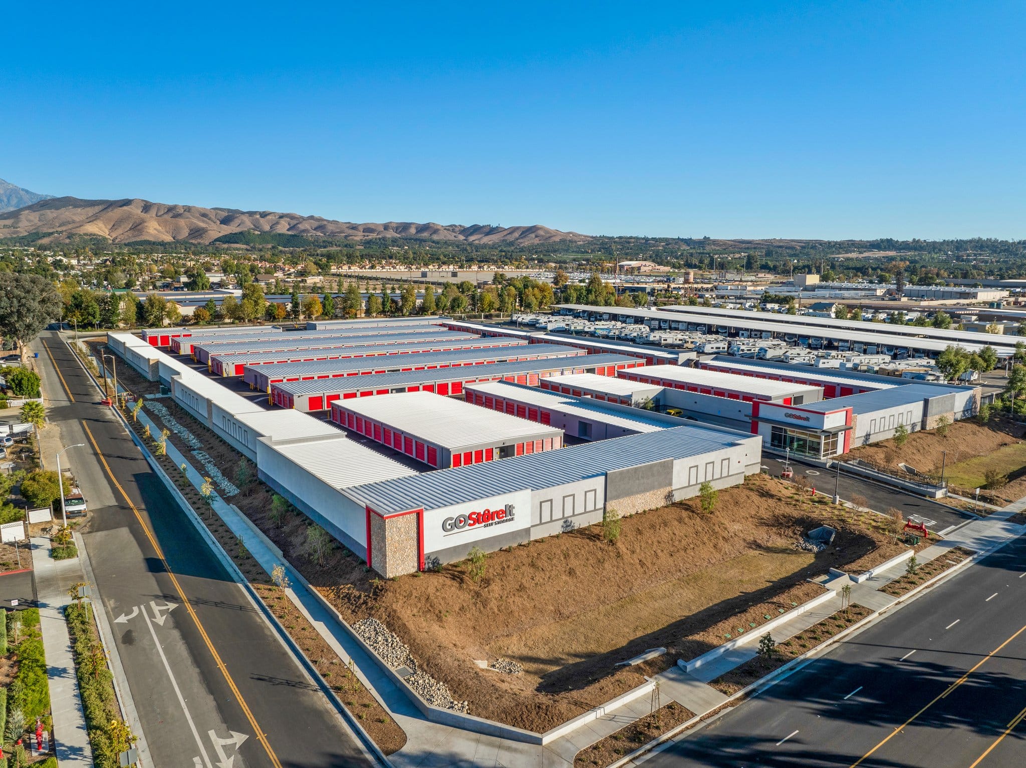 Aerial view of a large GoStoreIt self-storage facility with white and red buildings, surrounded by roads, parking areas, and nearby industrial buildings, with mountains in the background under a clear blue sky.